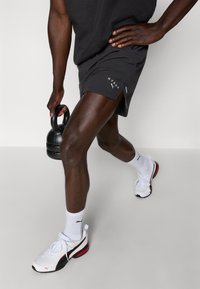 Melni šorti ar sānu iegriezumiem un baltu logo, baltas čības ar melniem un sarkaniem akcentiem, turēdams melnu kettlebellu uz neitrāla fona.