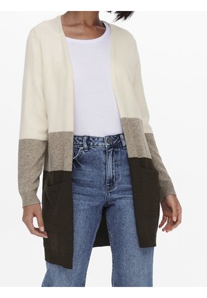 Frau trägt eine Farbblock-Strickjacke mit cremefarbenen, beigen und dunkelbraunen Abschnitten über einem weißen Hemd und blauen, hoch taillierten Jeans.