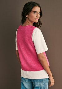 Rosa stickad väst med en mjuk textur, bärs över en vit t-shirt. Bakgrunden är en dämpad brunt, vilket framhäver plaggets färger.