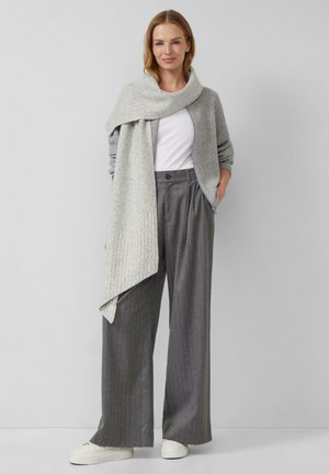 Femme debout portant un pantalon gris à jambes larges à rayures fines, un haut blanc, un cardigan gris zippé avec une écharpe assortie, et des baskets blanches.