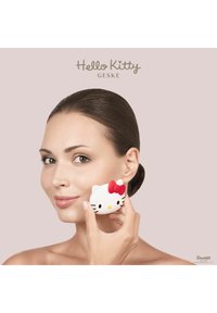 Hello Kitty skönhetssvamp i vitt med röd rosett och svarta morrhår, hålls mot kinden. Slät textur på en ljusrosa bakgrund.