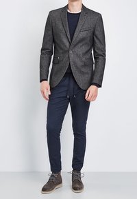 Męska ciemnoszara marynarka z wyciętymi klapami, dwoma guzikami i bocznymi kieszeniami, zestawiona z granatowymi spodniami na sznurku oraz brązowymi zamszowymi butami.