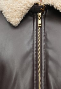 Bruine leren jas met een beige, gestructureerde shearlingkraag en een gouden rits met een hartvormige trekker.