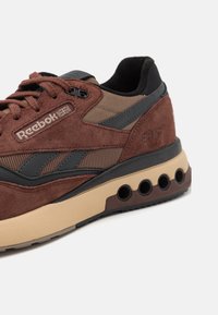 Bruna mockasiner i mocka med svarta och tan-mesh detaljer, gummisulor med cirkulära utskärningar och Reebok-logotyp på sidan.
