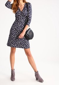 Marinblå omslagsklänning med vit blommönster, trekvartslånga ärmar, figursydd midja, kombinerad med en svart axelväska och grå ank stövlar.