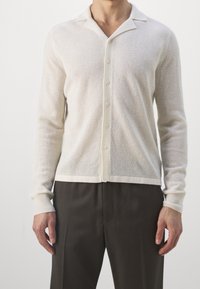 Cardigan leggero di colore crema, realizzato in maglia con un motivo testurizzato, maniche lunghe e colletto a punta, caratterizzato da una chiusura con bottoni sul davanti.
