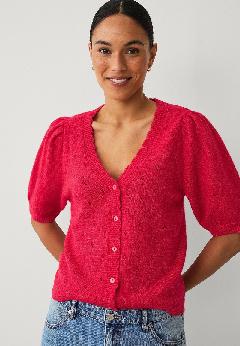 Žena se usmívá, nosí jasně červený propínací svetr s nafouknutými krátkými rukávy a světlé modré džíny, stojí na jednobarevném pozadí.