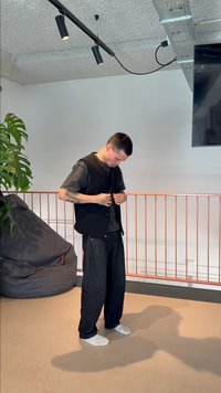 Mann med tatoveringer som knapper igjen svart vest, står barbeint på beige gulv nær en saccosekk og en inneplante i et lyst rom.