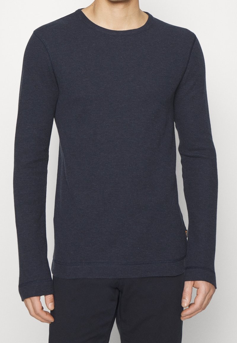 Homme portant une chemise unie bleu marine foncé à manches longues et un pantalon noir, debout, bras détendus le long du corps, sur fond blanc.