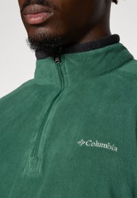 Green fleece pullover with a quarter-zip, features a black collar, smooth texture, and a white embroidered logo on the chest.