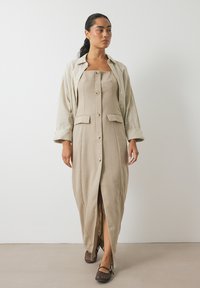 Beige linnedragt med lige snit, frontknapper og sidelommer, med en slidse op til låret. Matchende brune flade sko.