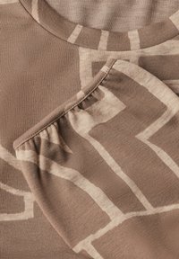 Brun mönstrad tyg med rundad halsringning och sammanklagad ärm. Designen inkluderar ljusbeige geometriska former. Mjuk textur.