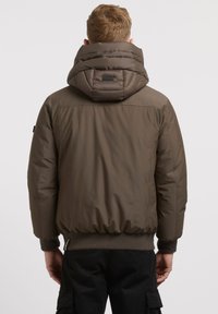 Veste matelassée vert olive avec capuche amovible, poignets et ourlet élastiques ; présente un col texturé et un logo au dos.
