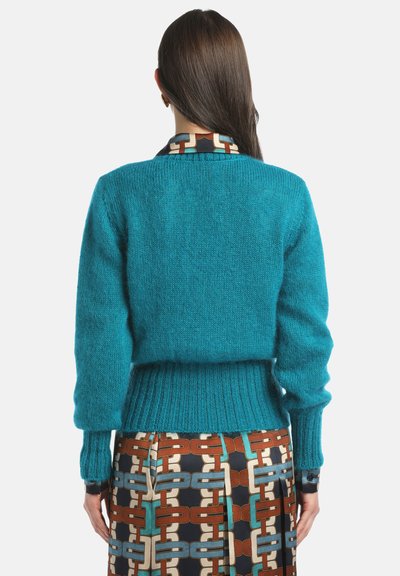 Türkiser gestrickter Pullover mit gerippten Bündchen und Saum; über einem gemusterten Rock getragen, der Verbindungen in Orange, Blau und Creme zeigt. Design mit hohem Kragen.