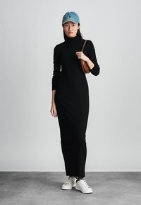 Melnas turtlenecks kleita ar garām piedurknēm, cieši pieguļoša līdz potītēm. Apvienota ar baltām kedām un zilu cepuri. Brūns crossbody soma.