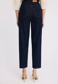 Kobieta w ciemnoniebieskich spodniach z wysokim stanem i beżowych szpilkach, stojąca tyłem na tle białej ściany.