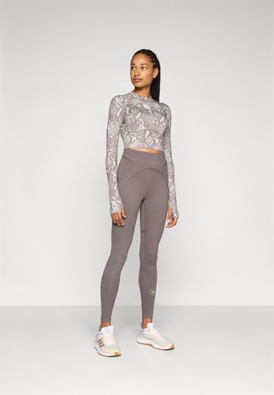 Woman standing wearing a long-sleeve cropped snake print top, high-waisted gray leggings, and white sneakers against a plain background.