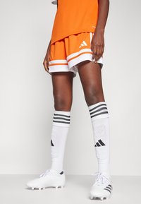 Oransje sportsdrakt kombinert med oransje shorts med hvite striper. Hvite sokker med svarte striper og svart logo, toppet med hvite fotballsko.