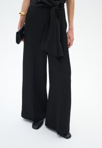 Pantalons larges noirs en tissu lisse. Détails d'un nœud noué à la taille. Accompagnés de chaussures plates noires et d'une pochette noire.