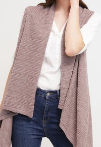 Gestrickter Schal in sanftem Mauve, mit einem strukturierten Muster, über einem weißen Hemd drapiert, das zu dunkelblauen Jeans passt. Keine Hardware oder Akzente sichtbar.