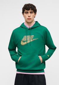 Νέος άνδρας που φοράει πράσινο φούτερ Nike με χρυσό λογότυπο και ροζ πουκάμισο από κάτω, τα χέρια στην μπροστινή τσέπη, ουδέτερο φόντο.