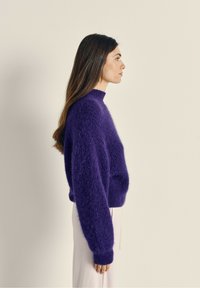 Violetti neulepaita, jossa on korkea kaulus, löysä malli ja pitkät ribattut hihat. Pehmeä, teksturoitu kangas. Puettu vaaleiden housujen päälle.