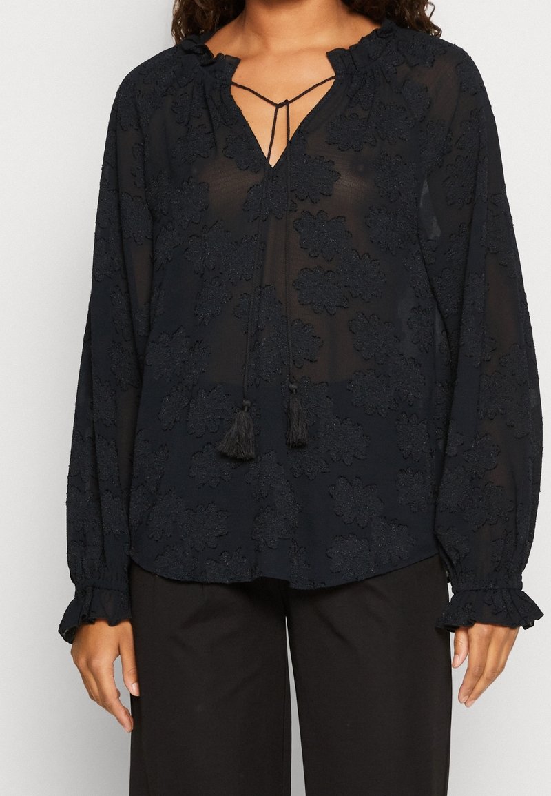 Blouse noire transparente à manches longues avec broderies florales, liens à pompons au niveau de l'encolure et poignets à volants, associée à un pantalon noir.