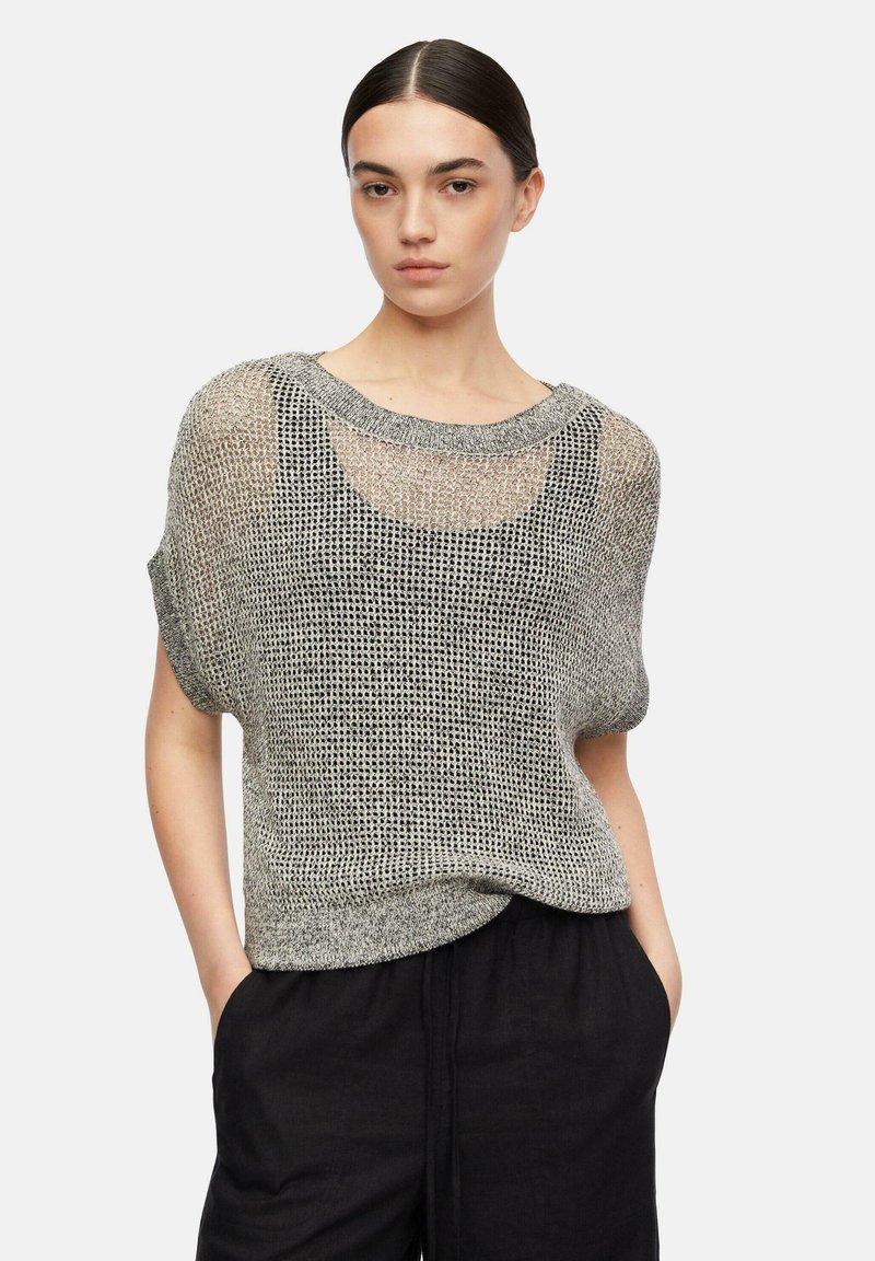 Junge Frau mit dunklem Haar in einem niedrigen Dutt, die ein lockeres graues Strickshirt mit kurzen Ärmeln über einem schwarzen Tanktop und lockere schwarze Hosen trägt, mit den Händen in den Taschen.