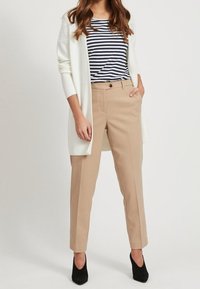 Beige byxor med skräddarsydd passform, i kombination med en marinblå och vit randig skjorta samt en lång vit kashmirtröja. Svarta högklackade skor fulländar looken.