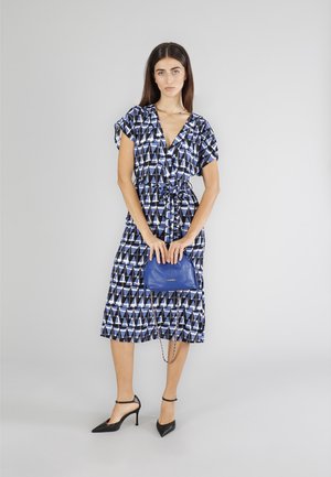 Abito multicolore con un motivo geometrico in blu, nero e bianco; vita cintrata, maniche corte e una gonna fluida. Una borsa blu lo completa.