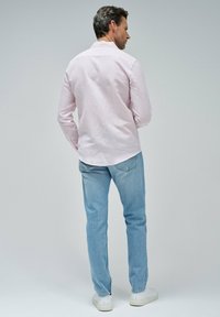 Camisa de manga larga rosa claro con cuello, combinada con jeans desgastados de color azul y zapatillas blancas, vista desde la parte trasera. Ajuste y estilo casual.