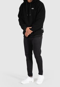 Svart fleece-hoodie med en framside-logga, i kombination med smala svarta byxor och vita sneakers. Har dragsko i huvan och ribbade muddar.