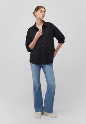 Schwarzes Hemd mit Knopfverschluss und Kragen, hochgekrempelte Ärmel, kombiniert mit hellblauen Schlagjeans und weißen Sneakers. Minimalistisches Design, schlichte Texturen.