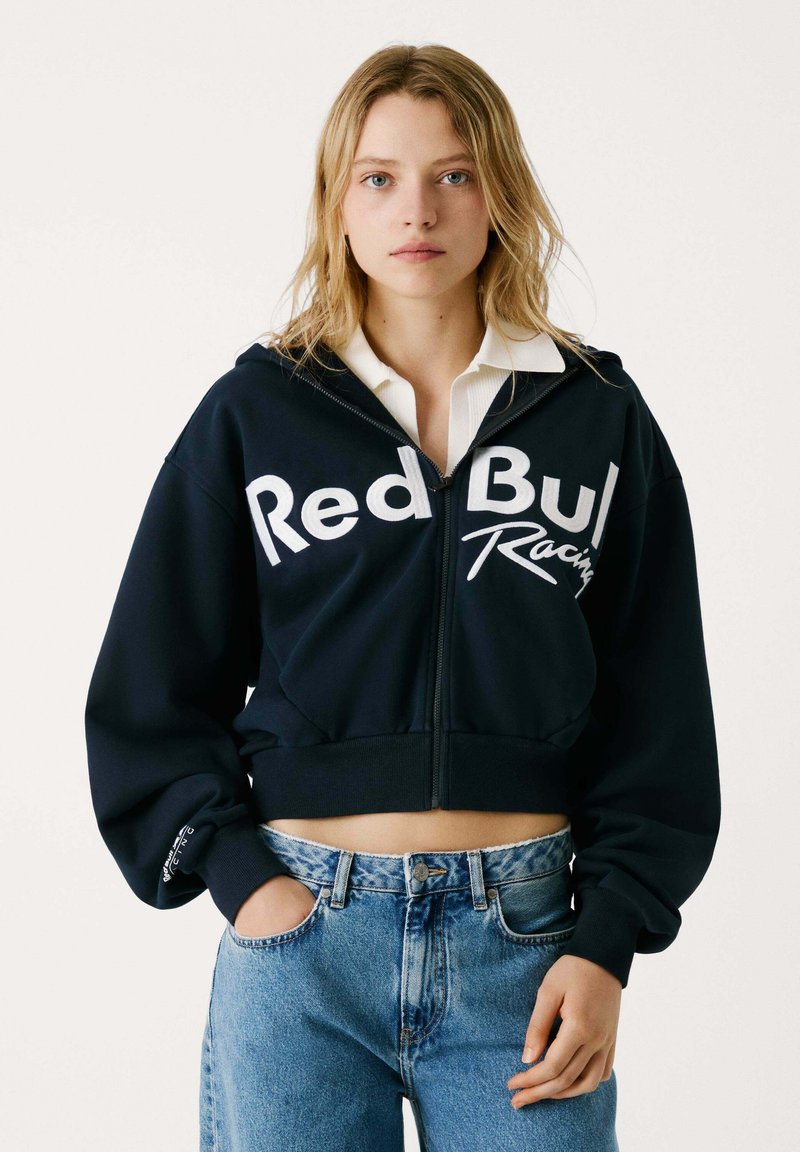 Mornarska skraćena dukserica s prednjim zipom, bijeli logo "Red Bull Racing", široki rukavi i kontrastni manšeti, u kombinaciji s visokim trapericama.