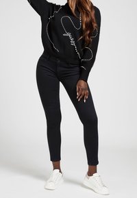 Schwarzer Pullover mit weißen Perlenakzenten, kombiniert mit figurbetonten dunklen Jeans und weißen Sneakers. Glatte Textur, schlankes Design und körpernahe Silhouette.
