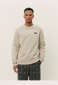 Beige Sweatshirt mit Rundhalsausschnitt und gerippten Bündchen, versehen mit einem kleinen schwarzen Logo-Patch auf der linken Brust. Untendrunter gemusterte Shorts.