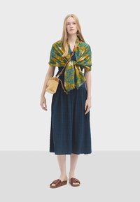 Langes, navyblaues Kleid aus strukturiertem Stoff, gemusterter Schal in Grün und Gelb sowie eine kleine beigen Tasche. Dunkelbraune Sandalen mit breiten Riemen.