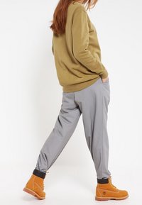 Felpa verde oliva, pantaloni grigi affusolati e stivali beige. I pantaloni hanno polsini neri. La felpa ha un design semplice senza loghi visibili.
