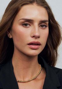 Guldhalsband med pärlor i rund och sfärisk form, bärs mot en svart topp och kompletterar subjektets ansiktsdrag. Subtil lyster.