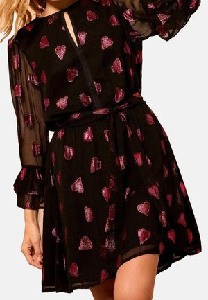 Schwarzes transparentes Kleid mit langen Ärmeln und roten glitzernden Herzmustern, mit Taillengürtel und kleinem Schlitz vorne am Ausschnitt.