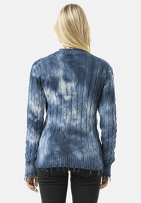 Pull en tricot bleu avec motif en cable, ourlet effiloché et effet dégradé tie-dye. Présente un col rond et des manches longues.