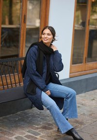 Marineblå quiltet frakke, sort tørklæde, sort top og flagrende lys blå jeans. Sidder på en bænk med en brostensbaggrund.