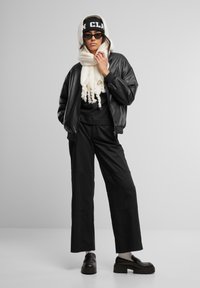 Giacca bomber in pelle nera sopra una maglietta nera e pantaloni larghi neri, abbinata a una sciarpa crema e un cappello nero. Mocassini neri chunky completano il look.