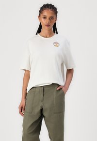 T-shirt crème à manches courtes avec un patch brodé rond au design coloré, associé à un pantalon large vert olive.
