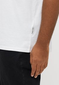 Λευκό βαμβακερό t-shirt με ευθεία γραμμή. Η πλαϊνή ραφή διαθέτει μια μικρή μαύρη ετικέτα με το κείμενο "PRAT COR". Φορώντας το με μαύρο παντελόνι.