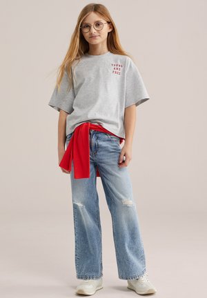 Tüdruk, kellel on pikk blondid juuksed ja prillid, kannab halli T-särki, millel on kiri "Young and Free", punast dressipluusi ümber vöökohal, rebenenud teksapükse ja tenniseid.