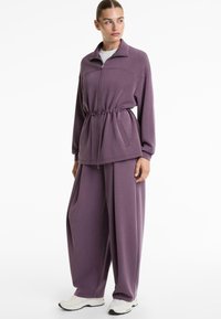 Lilla zip-up jakke med snor i taljen og lange ærmer, parret med løstsiddende lilla bukser. Hvide sneakers fuldender looket.