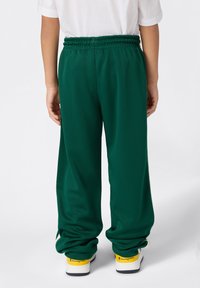 Ein Kind trägt weite grüneSweatpants und weiße Sneakers mit gelben Akzenten und steht mit dem Rücken zu einer schlichten weißen Wand.