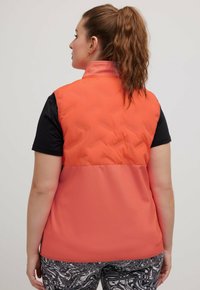 Orange puffer-väst med kviltad textur på den övre halvan, enfärgad nedre halva och hög krage, bärs över en svart skjorta.