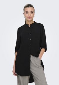 Camicia nera con colletto arrotondato, maniche lunghe arrotolate e un retro leggermente più lungo. Indossata con pantaloni grigi chiari.